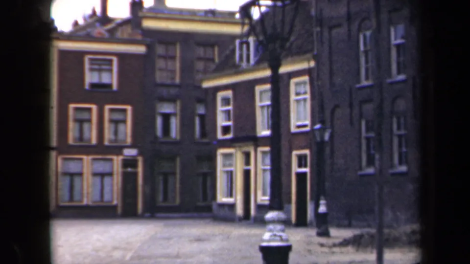 1946 Netherlands home-movie frame showing brick residential buildings with many windows, shutters, bare trees, and a foreground lamppost.
