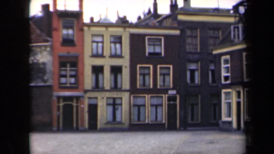 1946 Netherlands home-movie frame showing closer townhouse facades with red, ochre and dark masonry tones across a quiet street.