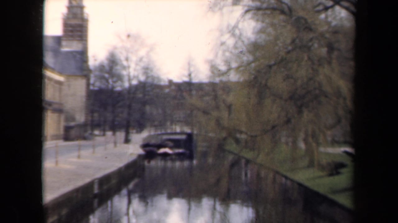 1946 Netherlands home-movie frame showing a canal, brick building, bare trees, and a dark car parked near the water.
