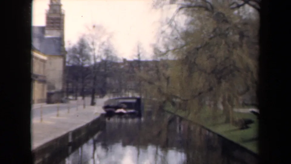 1946 Netherlands home-movie frame showing a canal, brick building, bare trees, and a dark car parked near the water.