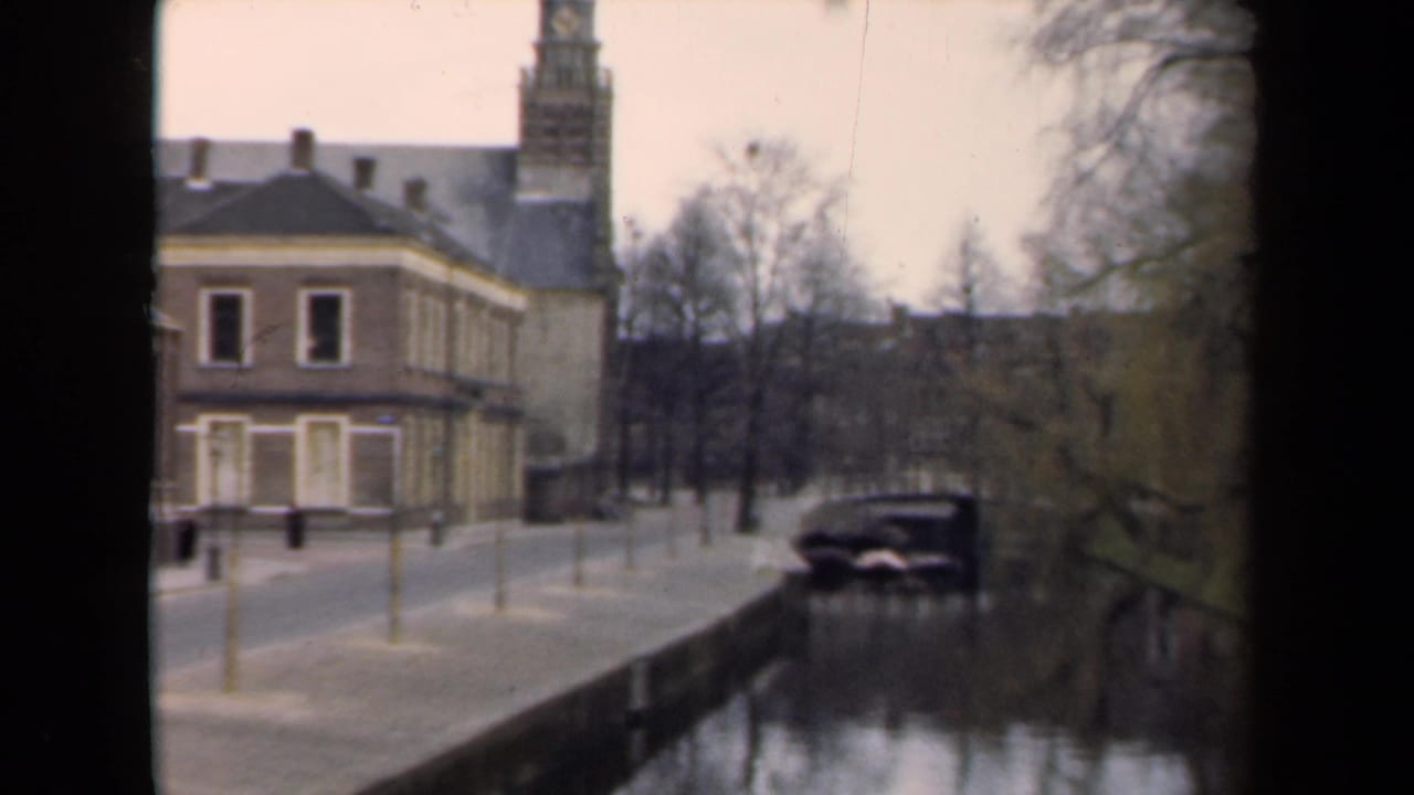 1946 Netherlands canal view with a brick building, tall tower, bare trees, yellow posts, and a parked car.
