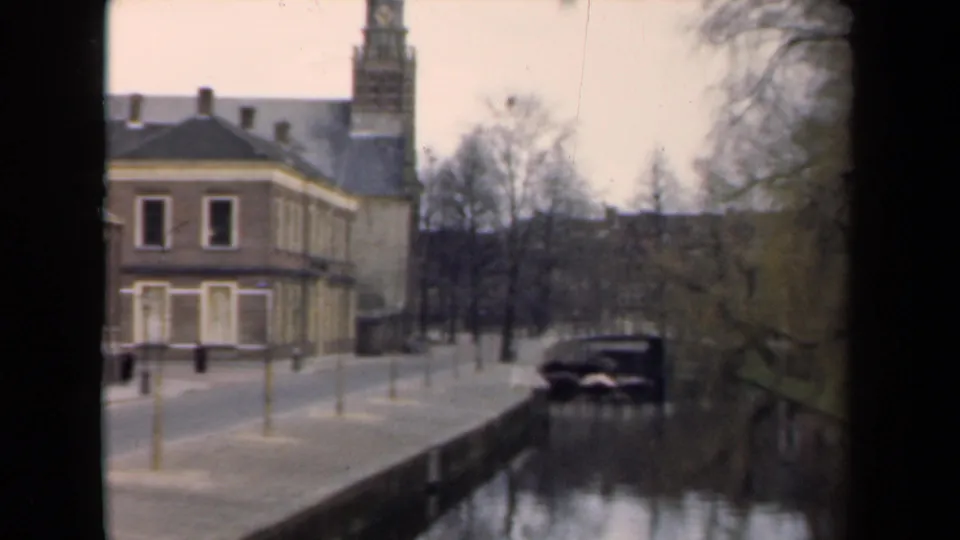 1946 Netherlands canal view with a brick building, tall tower, bare trees, yellow posts, and a parked car.