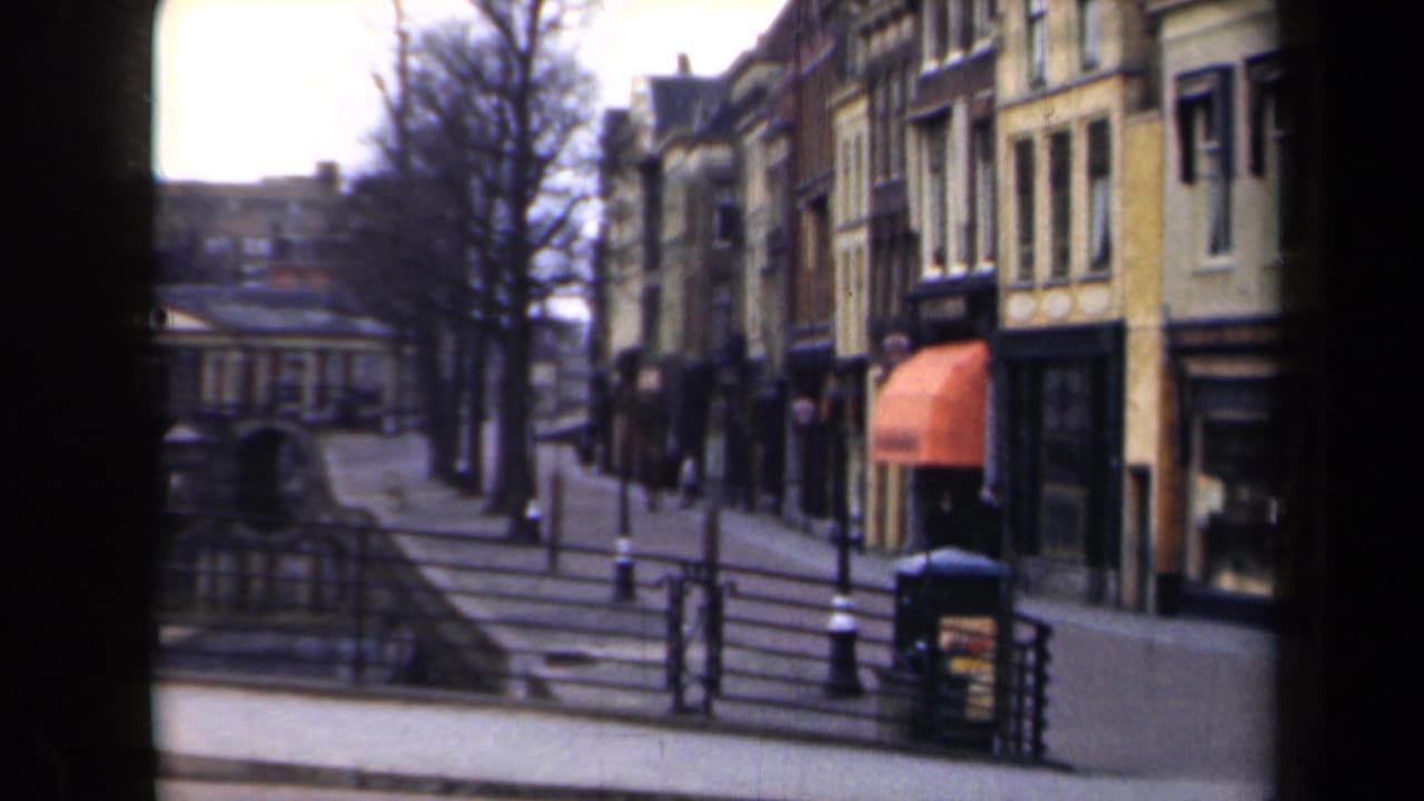 1946 Netherlands home-movie frame showing a canal-side promenade with steps, railings, bare trees, shopfronts, and an orange awning.