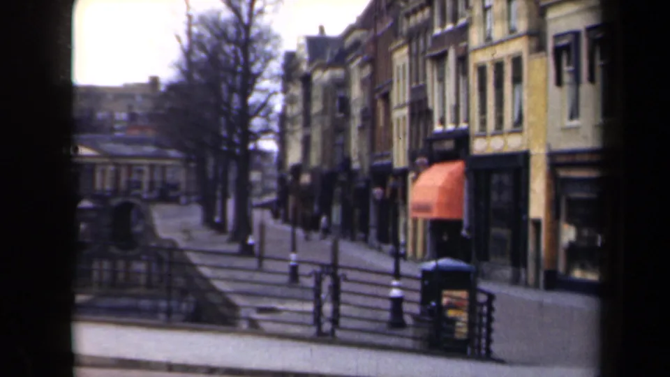 1946 Netherlands home-movie frame showing a canal-side promenade with steps, railings, bare trees, shopfronts, and an orange awning.