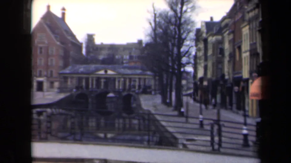 1946 Netherlands frame showing a canal-side street, red brick building, arched structure, bare trees, railings, and shopfronts.