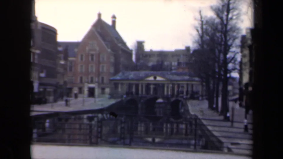 1946 Netherlands home-movie frame with a canal bridge, red-brick gabled building, columned waterside structure, bare trees, and small pedestrian figures.