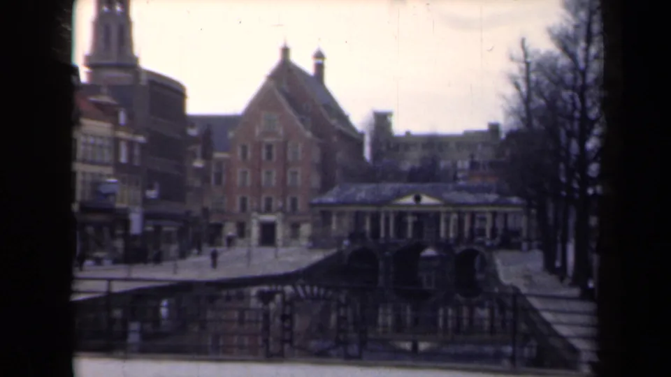 1946 Netherlands 8mm frame showing a canal-side bridge, brick gabled architecture, railings, bare trees, and pedestrians in a quiet town setting.