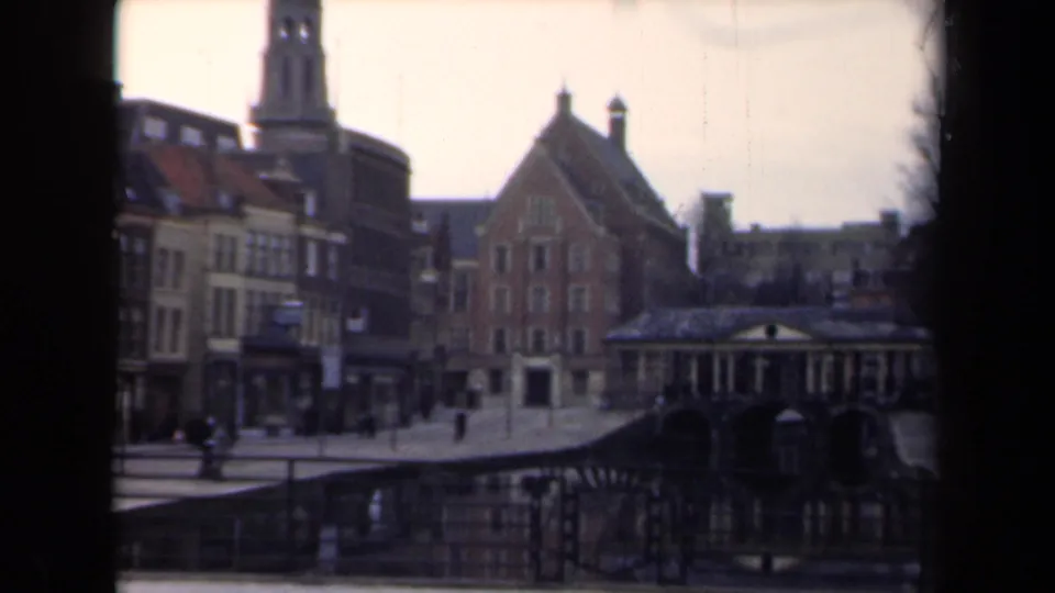 1946 Netherlands 8mm frame with a canal bridge, brick gabled building, partial tower, waterside railings, bare trees, and distant pedestrians.