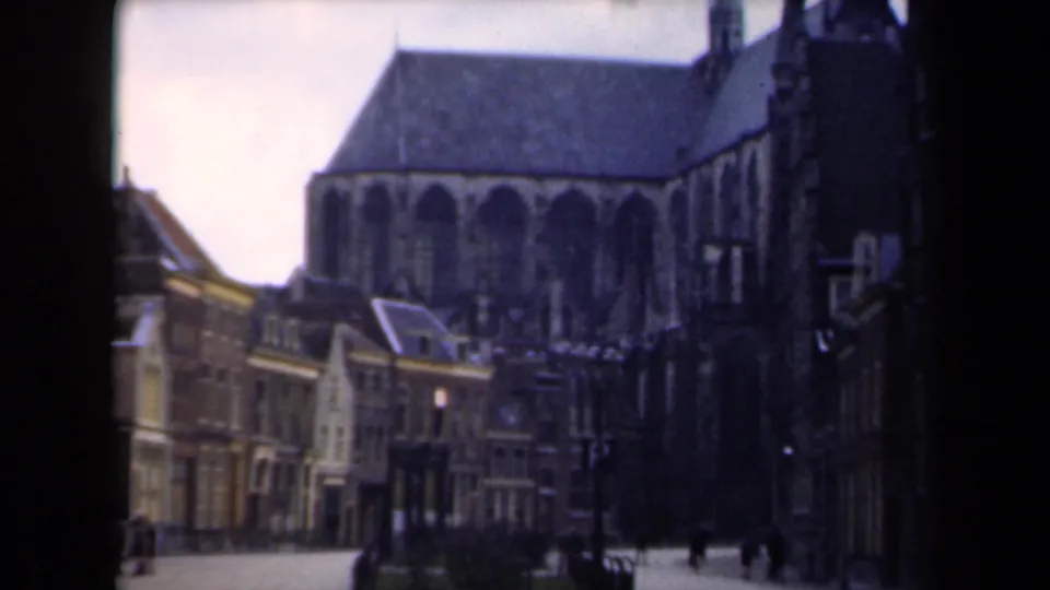 Wider 1946 Netherlands home-movie view of a church exterior, adjoining town buildings, paved square, and distant pedestrians.