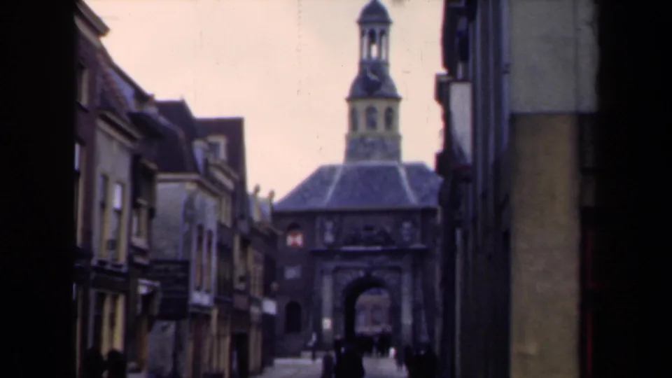 Faded 8mm frame of a 1946 Dutch street, showing pedestrians, older facades, and a stone gatehouse with tower.