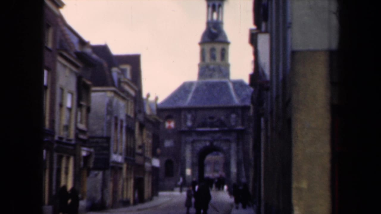 1946 Netherlands home-movie frame showing a narrow street, traditional buildings, a gate tower, and distant pedestrians.