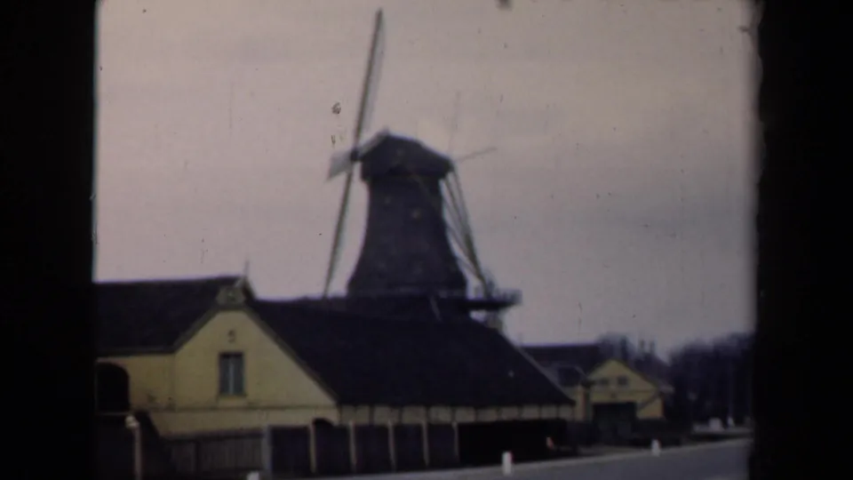 1946 Netherlands home-movie frame showing a windmill, yellow-toned building, dark roofs, and quiet roadside setting.
