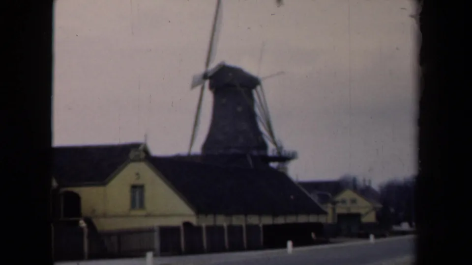 1946 Netherlands home-movie frame with a windmill rising over traditional low buildings beside a quiet road.