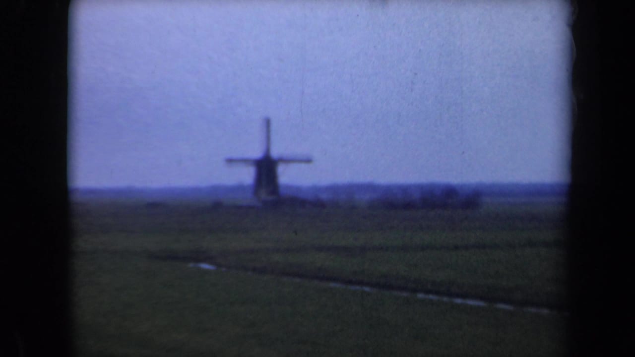 Distant windmill across a flat green field under a pale blue sky.