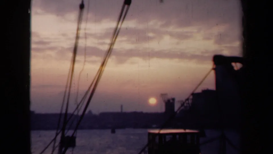 Archival 8mm Netherlands harbor sunset with foreground rigging, a dark vessel edge, calm water, and distant waterfront silhouettes.