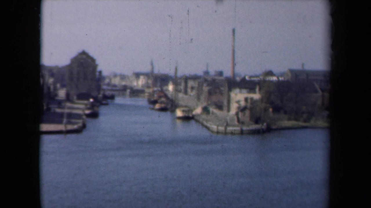 1946 Netherlands home-movie frame showing a canal or marina edge with buildings, moored boats, and a tall chimney.