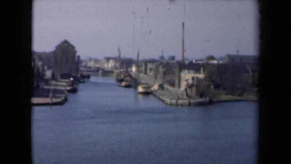 1946 Netherlands home-movie frame showing a canal or marina edge with buildings, moored boats, and a tall chimney.