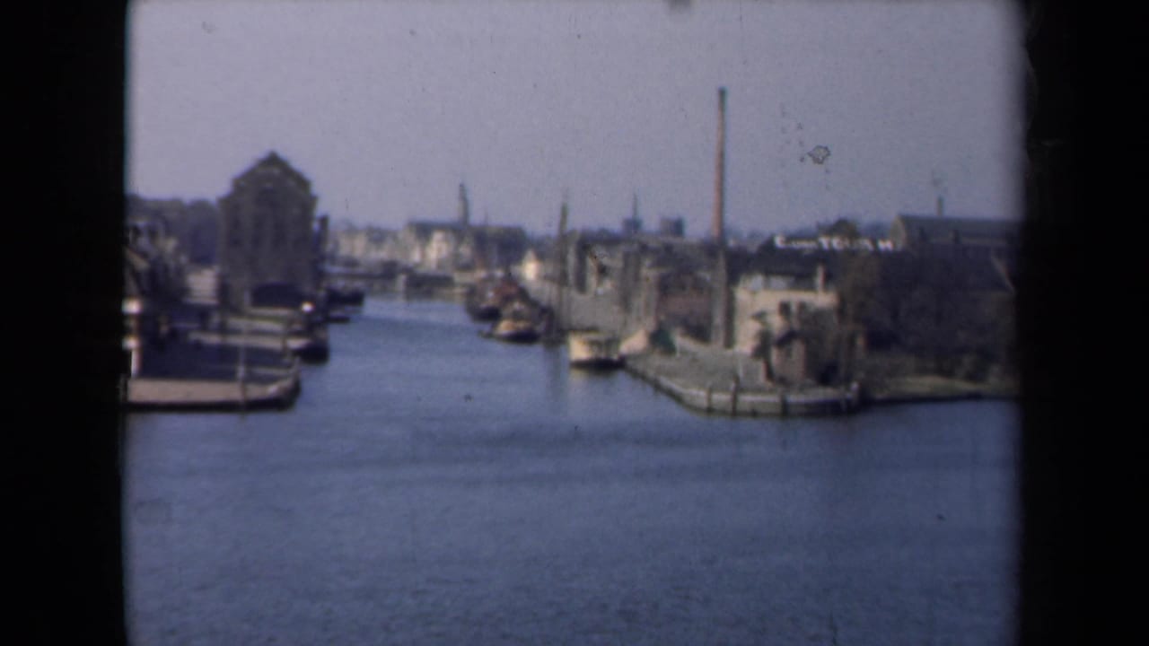 1946 Netherlands 8mm frame showing a canal lined with boats, old waterfront buildings, and a distant industrial chimney.