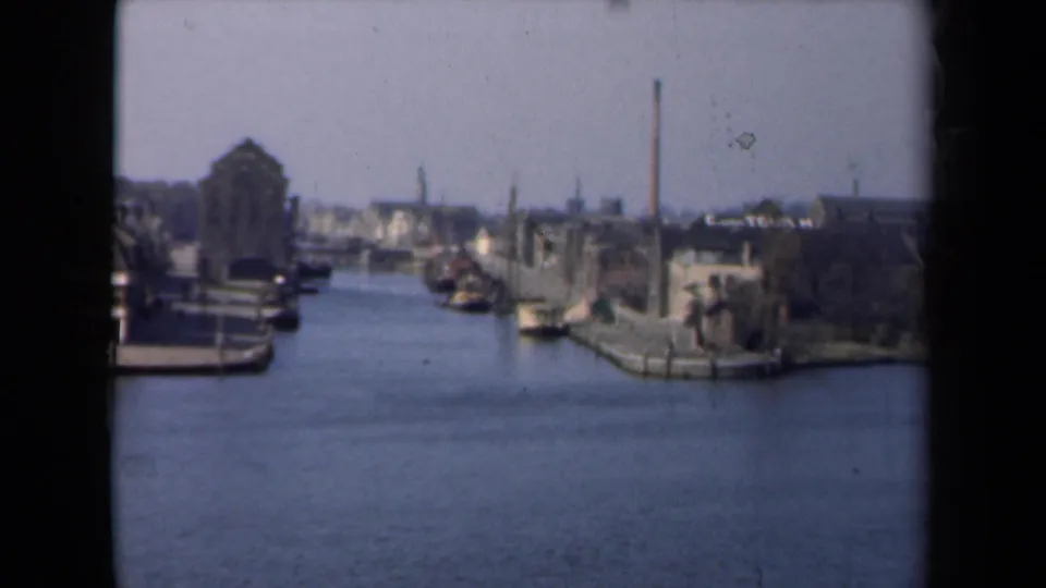 1946 Netherlands 8mm frame showing a canal lined with boats, old waterfront buildings, and a distant industrial chimney.
