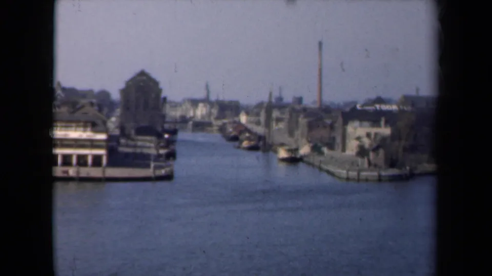 1946 Netherlands canal view from a moving boat, with moored vessels, quayside buildings, chimneys, and soft distant detail.