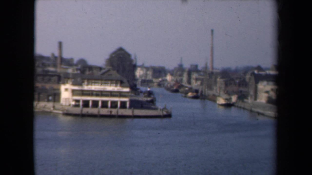 1946 Netherlands 8mm view of a canal with a docked riverboat, quayside buildings, smokestacks, and a hazy industrial skyline.