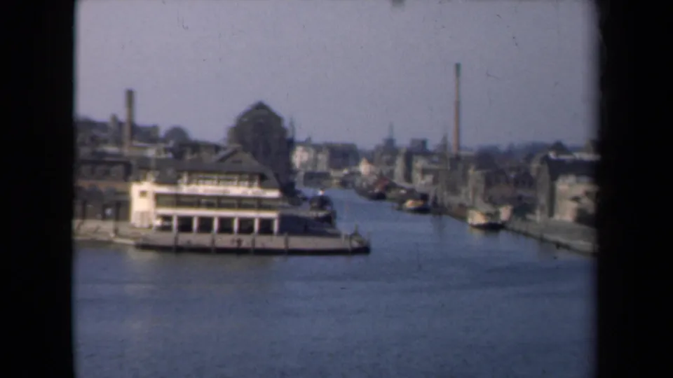 1946 Netherlands 8mm view of a canal with a docked riverboat, quayside buildings, smokestacks, and a hazy industrial skyline.