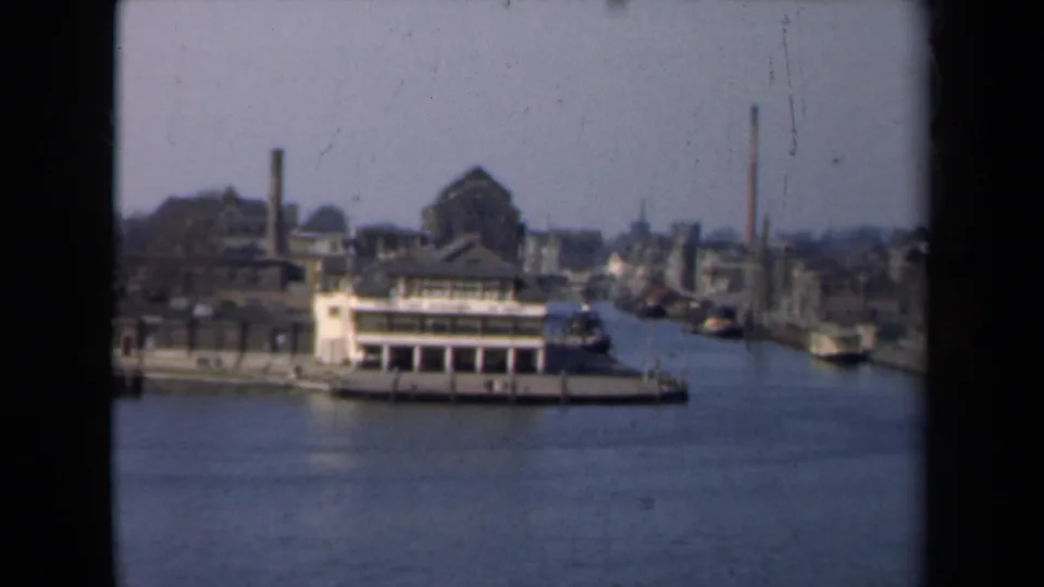 1946 Netherlands canal scene showing a moored riverboat, low waterfront structures, smokestacks, and soft 8mm film texture.