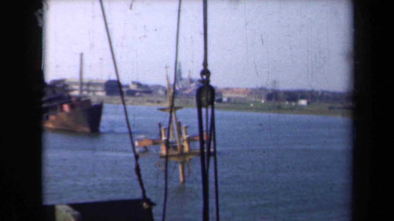 Boat-level 1946 Netherlands waterway view with rigging in the foreground, vessels on the river, and low industrial buildings along the far bank.