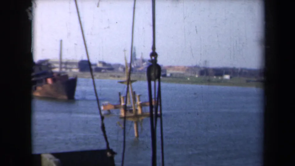 Boat-level 1946 Netherlands waterway view with rigging in the foreground, vessels on the river, and low industrial buildings along the far bank.