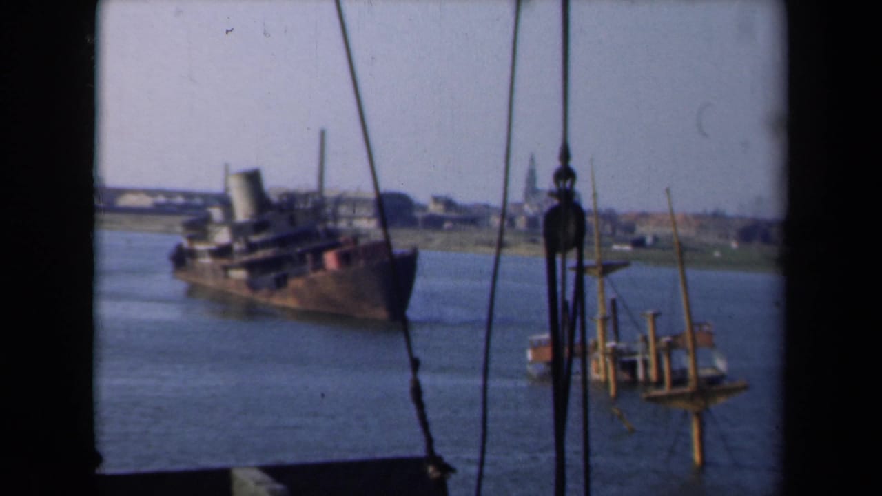 A damaged vessel passes near small waterway structures, with a distant Dutch skyline and steeple visible beyond the shore.