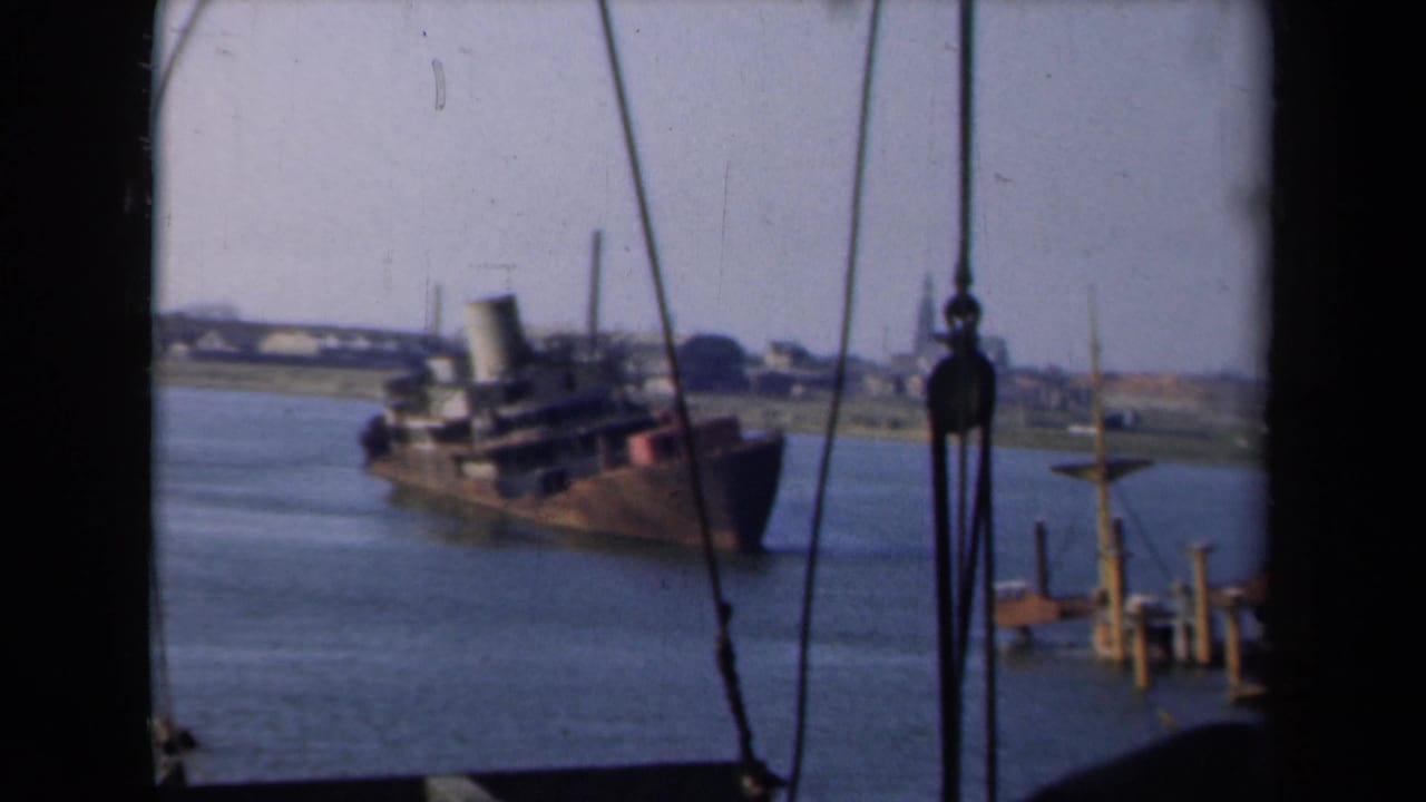 The ship appears in profile in the Netherlands waterway, framed by rigging with low shoreline buildings in the background.