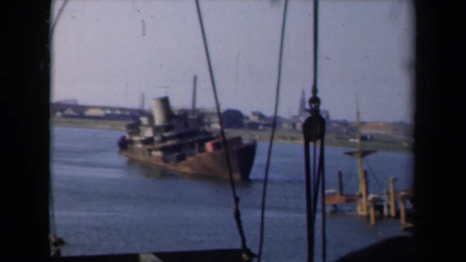 The ship appears in profile in the Netherlands waterway, framed by rigging with low shoreline buildings in the background.