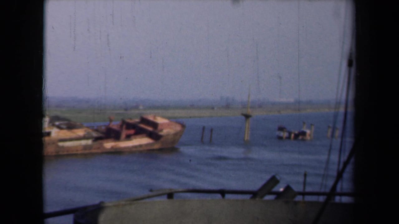 Grounded vessel section near pilings in a broad shallow waterway under a hazy sky.