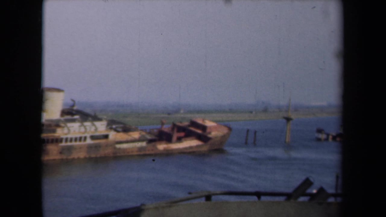 1946 Netherlands 8mm frame showing the listing steamer closer in view, with pilings and flat shoreline beyond the water.