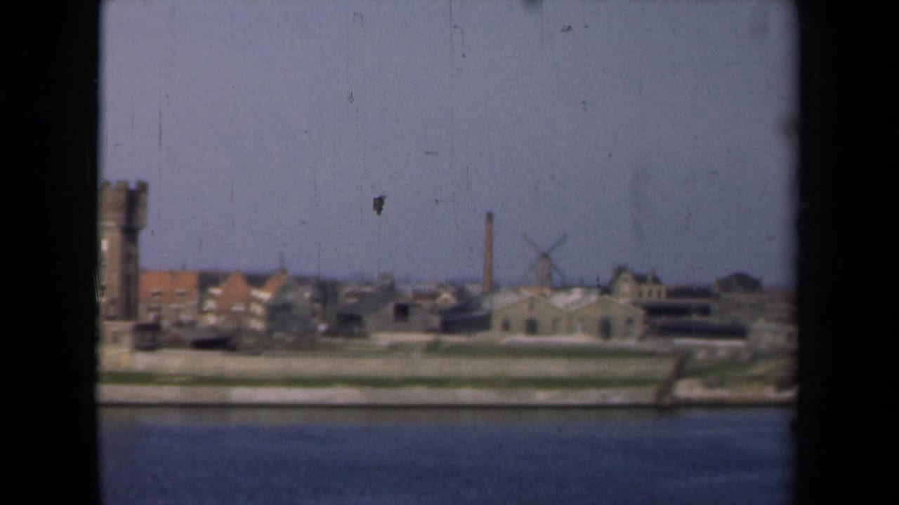 1946 Netherlands 8mm frame showing a waterside townscape with a tower, low buildings, a chimney, and a windmill under a hazy sky.