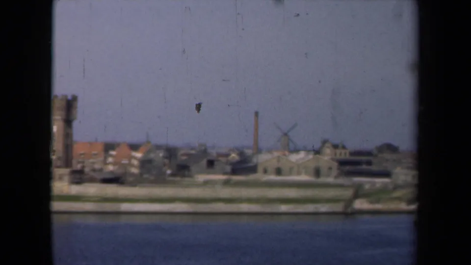 1946 Netherlands 8mm frame showing a waterside townscape with a tower, low buildings, a chimney, and a windmill under a hazy sky.