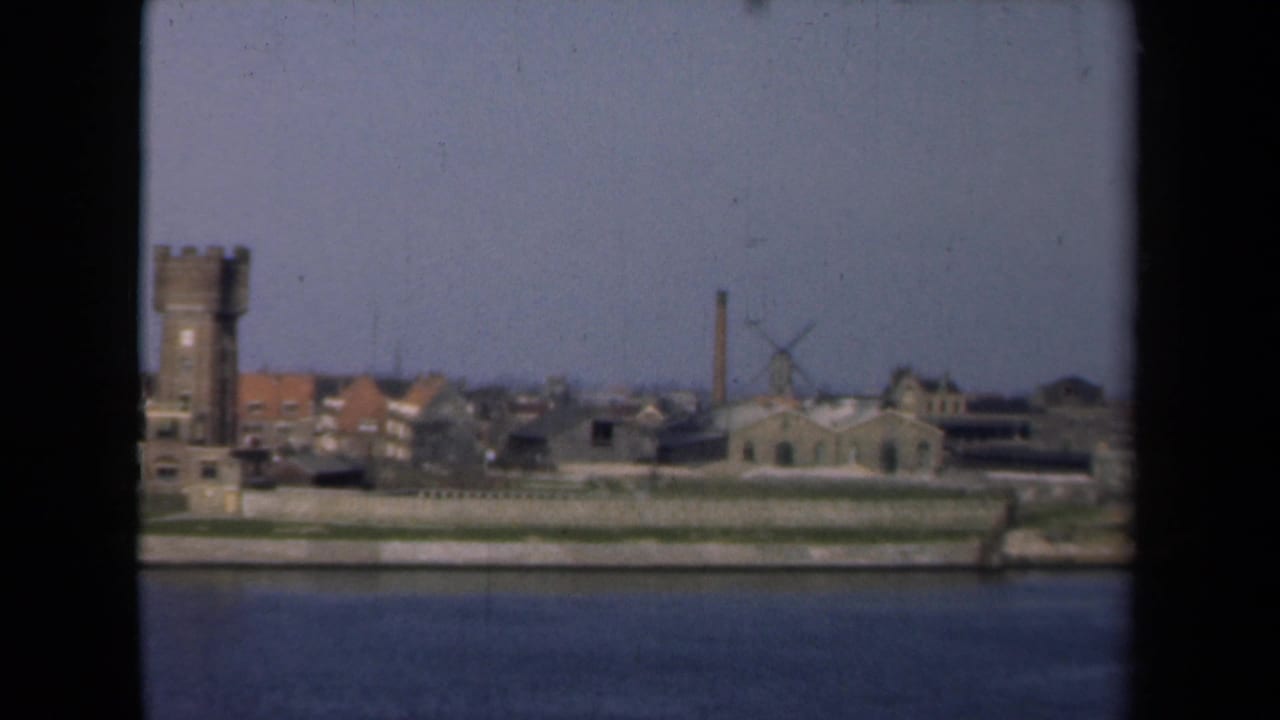 Riverfront townscape with stone tower, buildings, windmill, chimney, and blue sky.