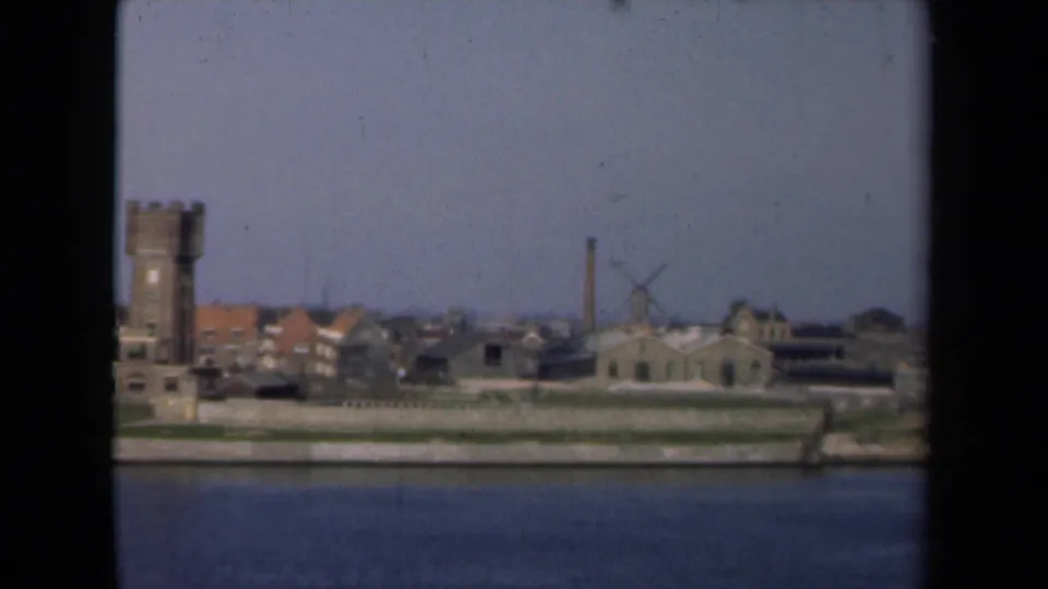 1946 Netherlands river view showing a waterfront townscape with a stone tower, windmill, chimney, and low buildings beyond calm water.