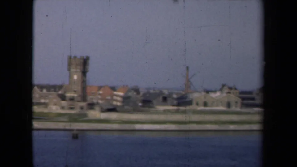 Soft 8mm frame from a 1946 Netherlands river passage, showing a tower, waterfront structures, windmill, chimney, and broad sky.