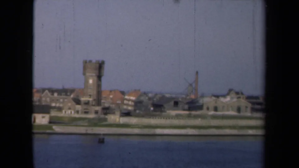 End frame of the 1946 Netherlands riverfront view, with calm water, a stone tower, low buildings, a windmill, and chimney under pale sky.