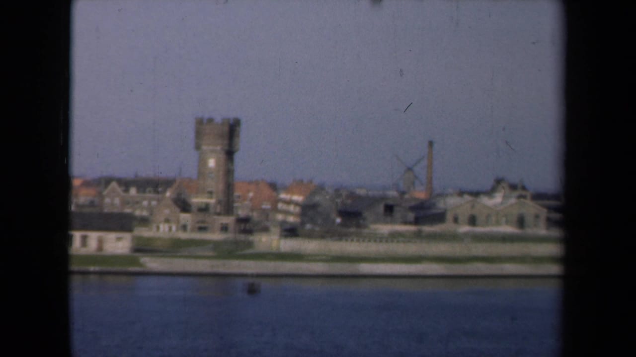 1946 Netherlands 8mm view of a waterfront town with a masonry tower, red-roofed buildings, and a windmill seen from across the water.