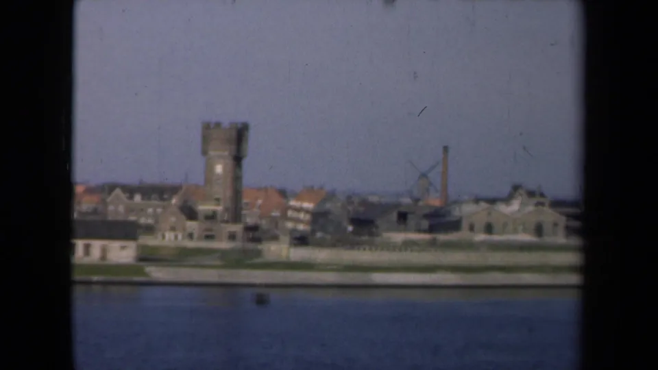 1946 Netherlands 8mm view of a waterfront town with a masonry tower, red-roofed buildings, and a windmill seen from across the water.