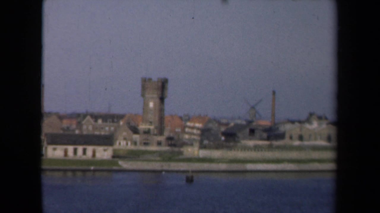 Similar waterfront view with tower, windmill, houses, and broad water in the foreground.