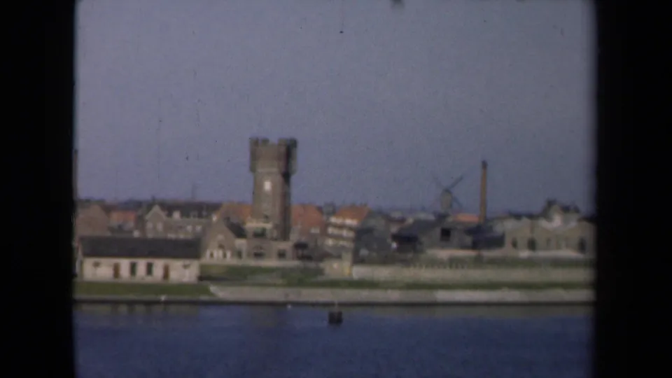 Dutch waterfront in 1946 with a tall tower, windmill, clustered buildings, and calm water, framed by dark 8mm film borders.