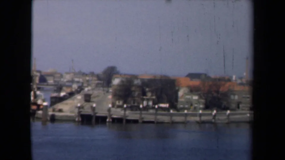 1946 Netherlands waterfront quay with piers, moored boats, bare trees, and low town buildings viewed from a boat.