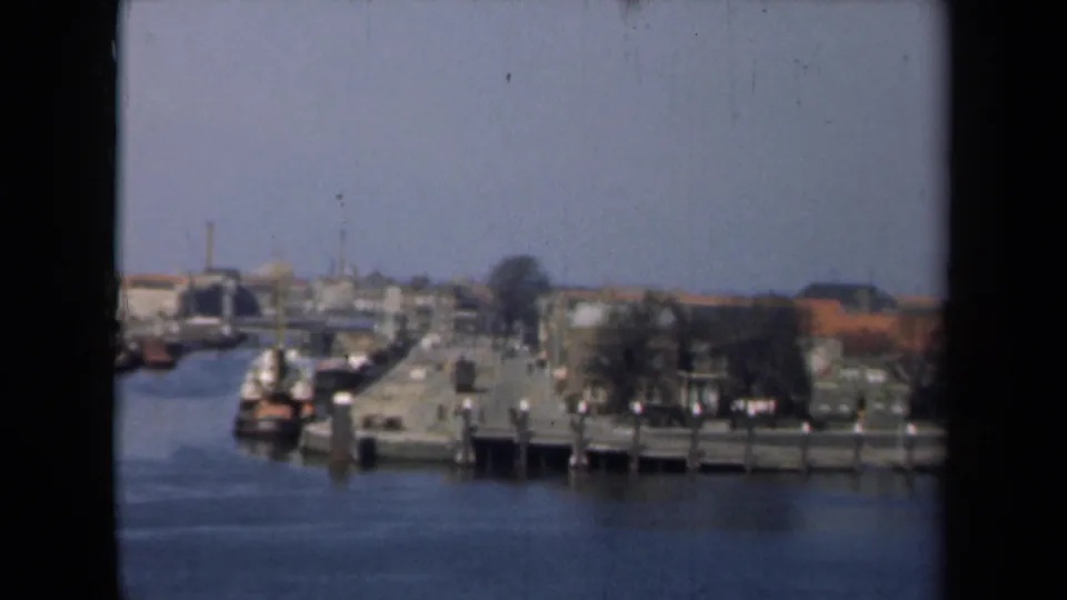 Dutch harborfront in 1946 showing piers, small boats, bare trees, and low buildings across the water in soft 8mm color.