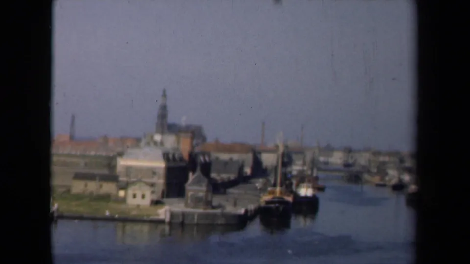 1946 Netherlands harbor view showing calm canal water, docked boats, waterfront structures, and a church steeple above the town.