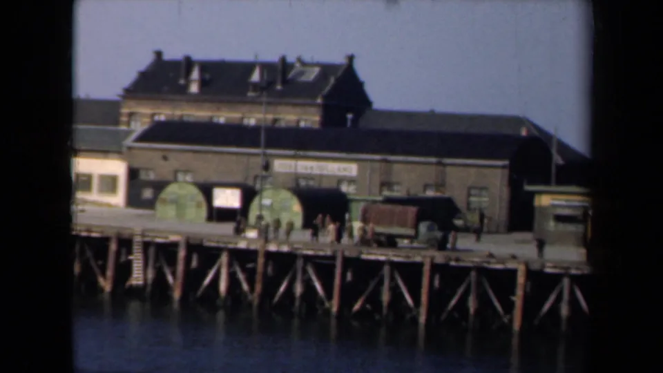 1946 Netherlands dockside scene with a waterfront warehouse, visible signage, workers, carts, and wooden pier supports over the water.