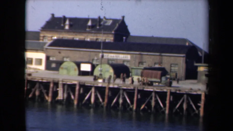 1946 Netherlands 8mm frame of a dockside warehouse with signage, workers on the pier, carts, and dark water below.
