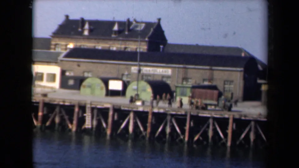 1946 Netherlands dockside view with an industrial building, green tanks, a military-style truck, and small figures on a timber pier.